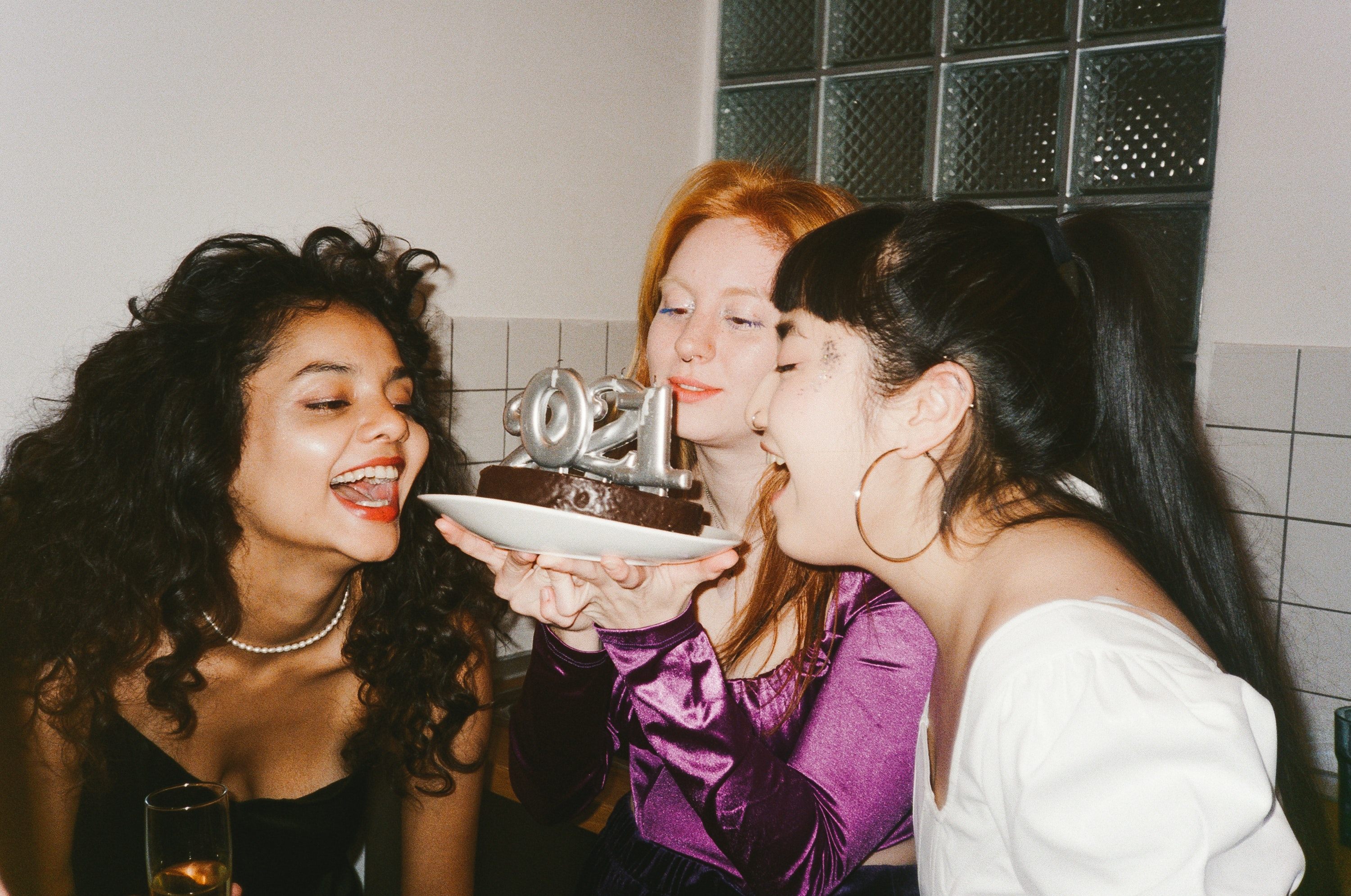 Three girls, indoors, having a good time, holding a cake with candles in it.